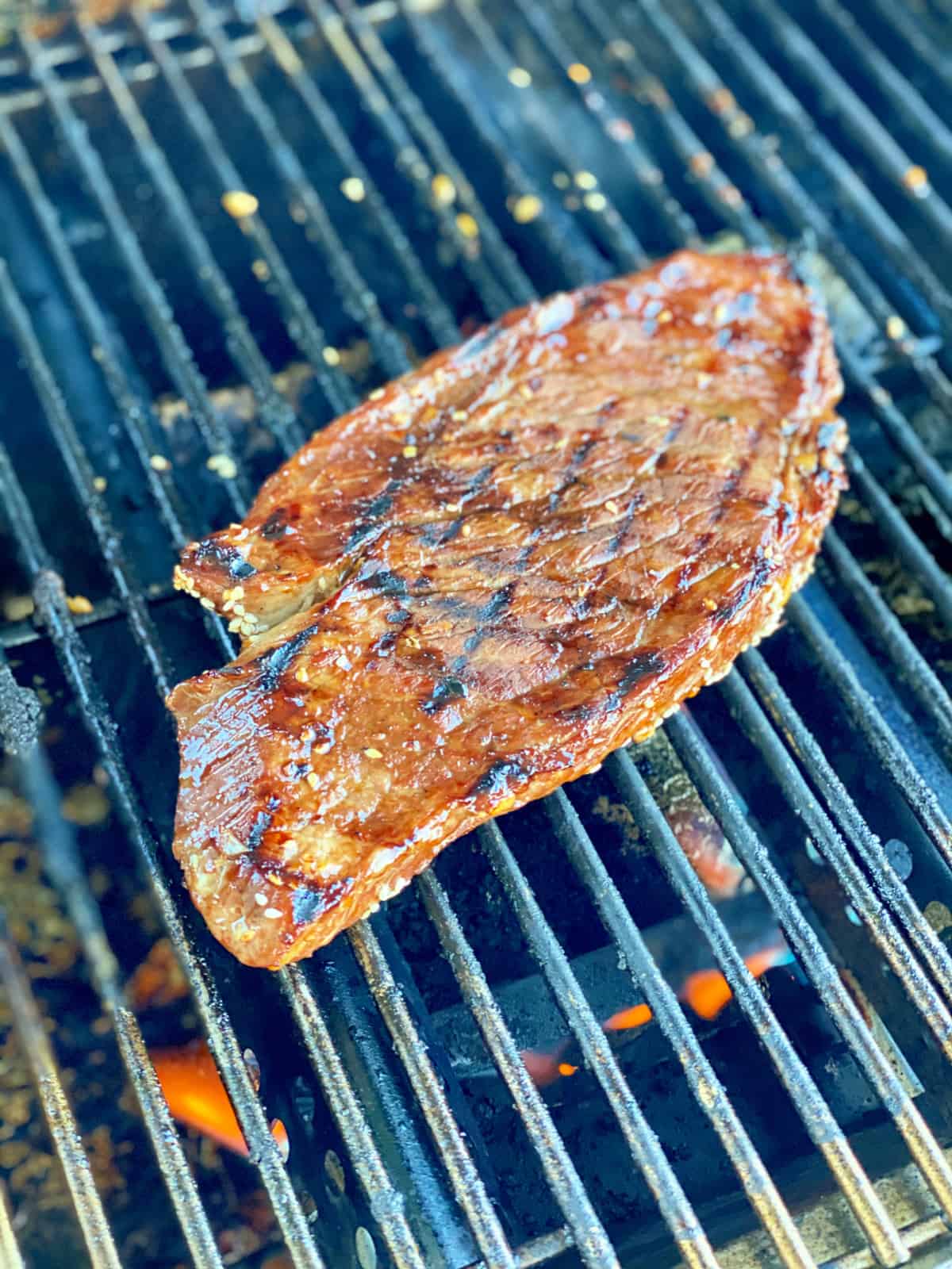 Large london broil steak with grill marks on top of a grill with fire underneath.