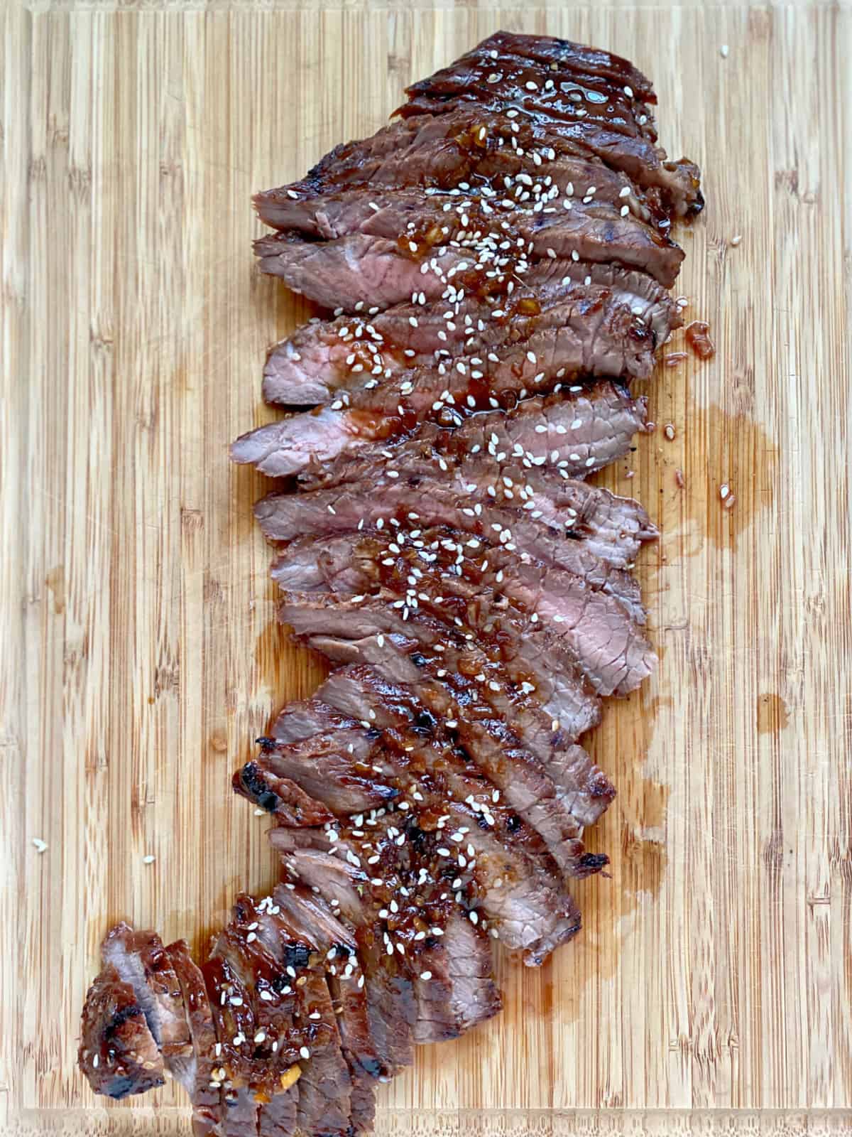 Top view of a wood cutting board with sliced steak topped with sauce and sesame seeds.