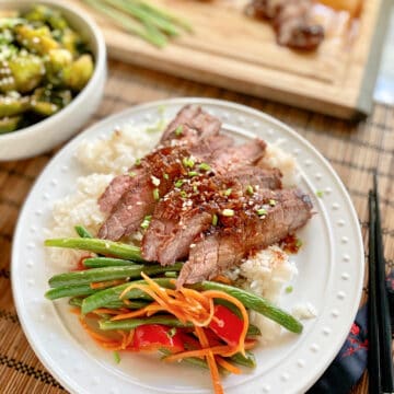 White plate filled with rice, green beans, carrots, bell peppers, and sliced steak with steak on a cutting board in the background.