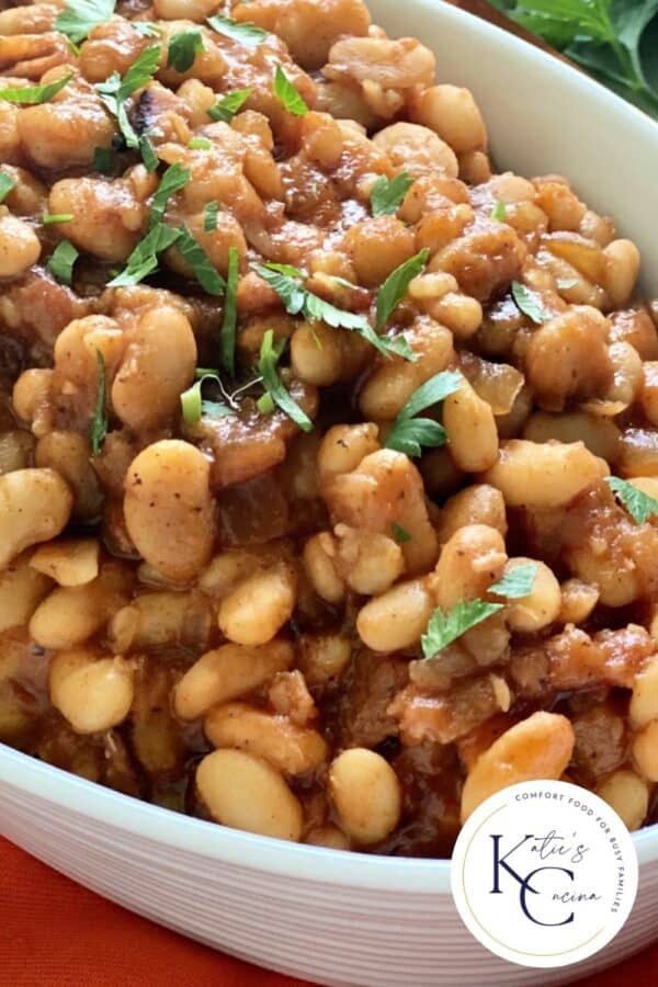 Close up of baked beans in a white oval bowl with logo on right corner.