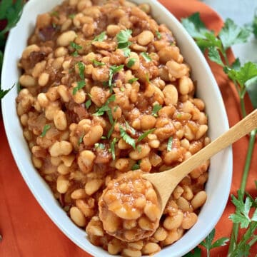 Oval white bowl filled with baked beans with a wooden spoon in the bowl sitting on an orange cloth.