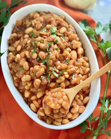 Oval white bowl filled with baked beans with a wooden spoon in the bowl sitting on an orange cloth.