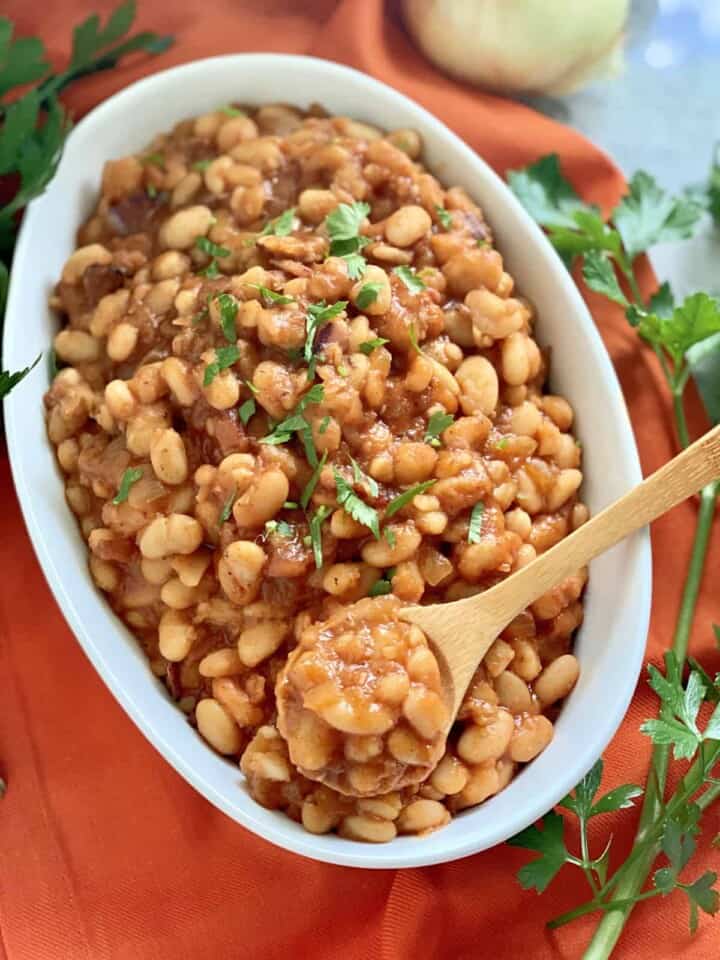 Oval white bowl filled with baked beans with a wooden spoon in the bowl sitting on an orange cloth.