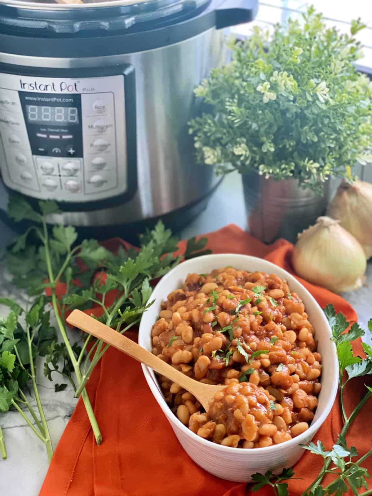 Oval bowl of baked beans on cloth with herbs, onions, and Instant Pot in backgound.