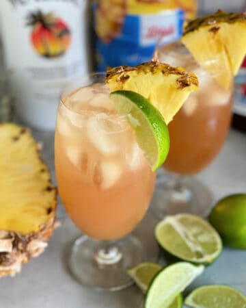 Two glasses of peach colored cocktails with pineapples and limes.