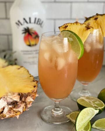 Two glasses filled with peach colored juice with limes and pineapple on counter.