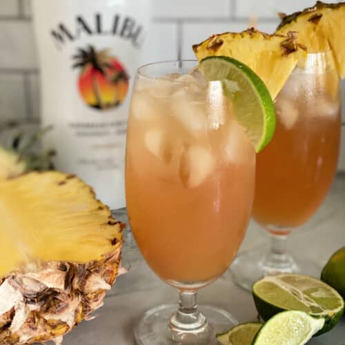 Two glasses filled with peach colored juice with limes and pineapple on counter.