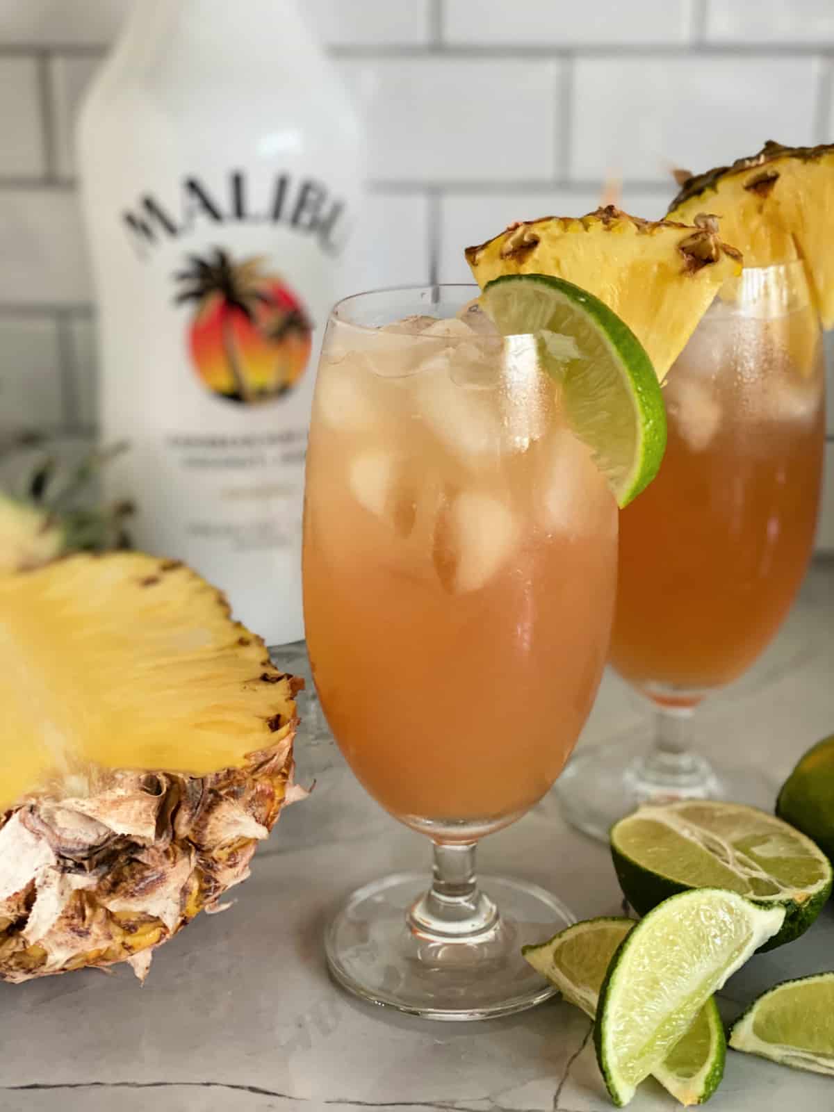 Two glasses filled with peach colored juice with limes and pineapple on counter.