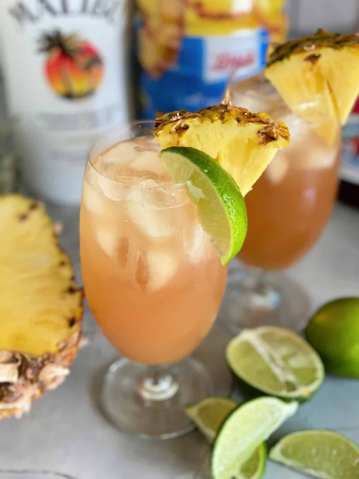 Two glasses of peach colored cocktails with pineapples and limes.