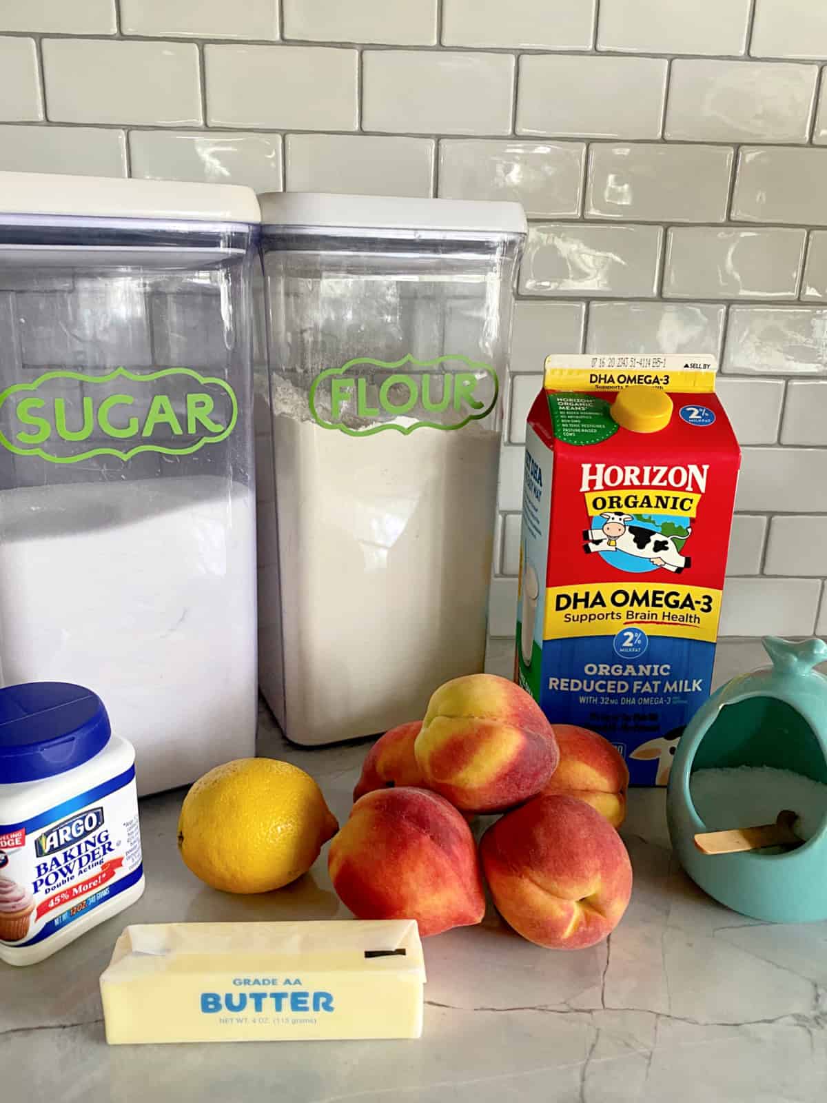 Ingredients on counter: Sugar, flour, baking powder, butter, lemon, peaches, milk, and salt.