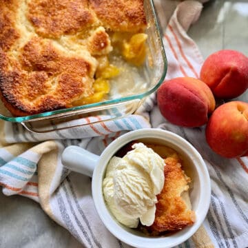 Top view of a white crock with ice cream and peach cobbler, a glass baking dish with peach cobbler, and fresh peaches next to it.