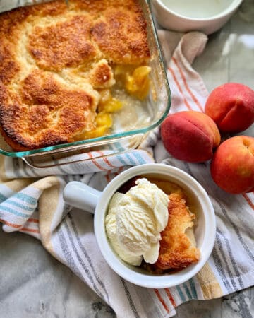 Top view of a white crock with ice cream and peach cobbler, a glass baking dish with peach cobbler, and fresh peaches next to it.
