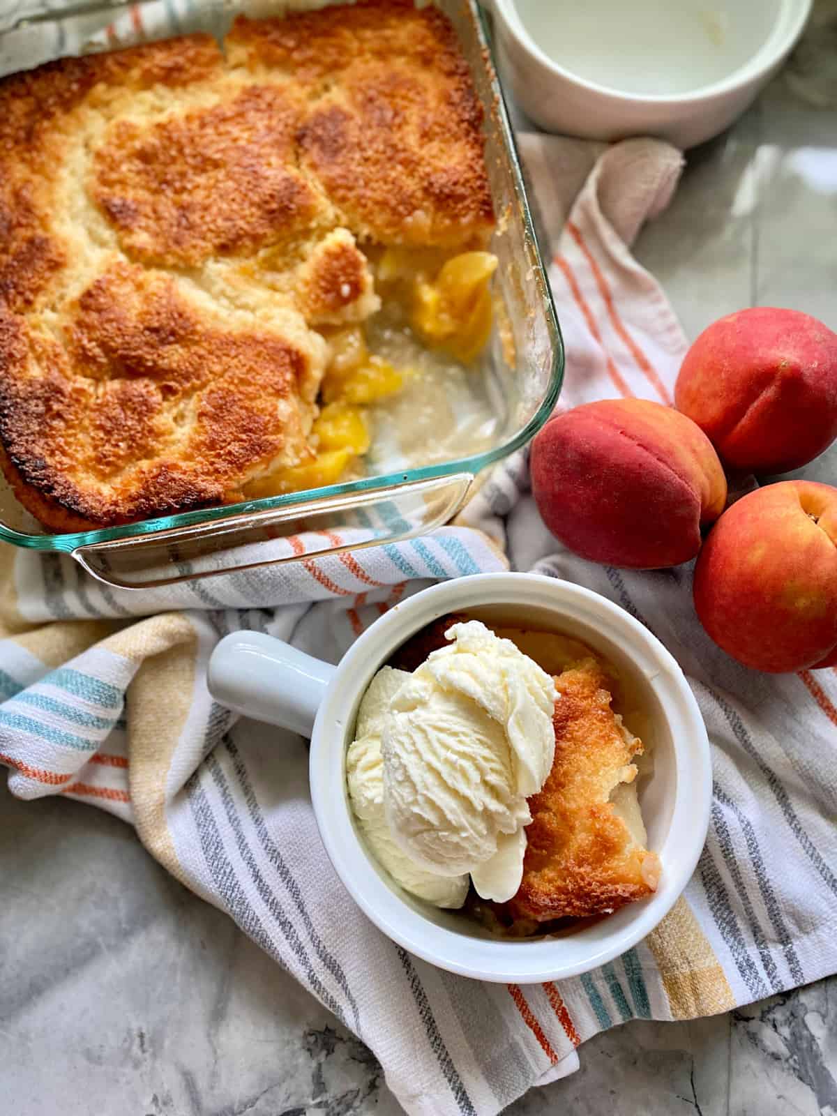 Top view of a white crock with ice cream and peach cobbler, a glass baking dish with peach cobbler, and fresh peaches next to it.
