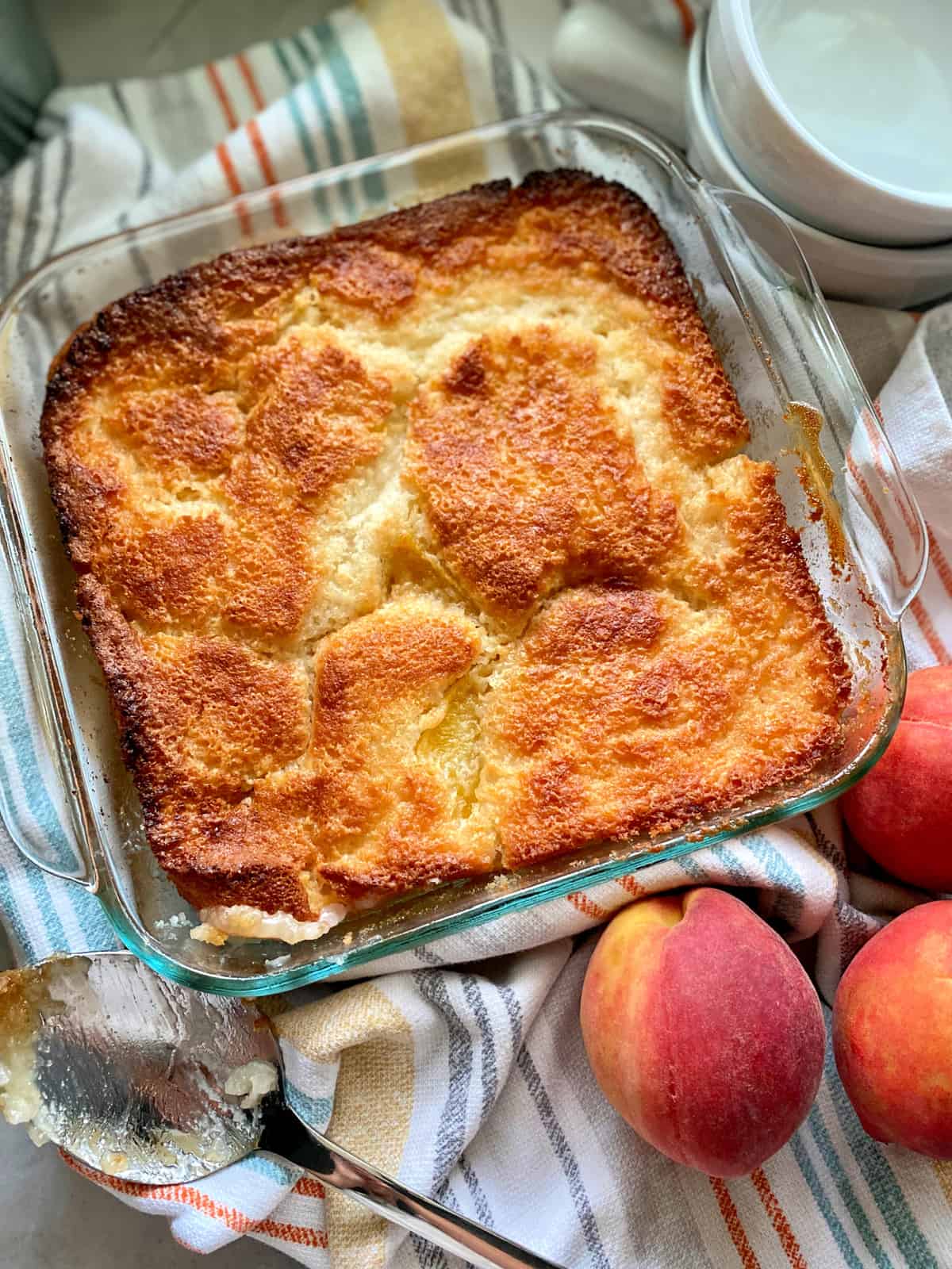 Top view of a square glass baking dish filled with peach cobbler with a spoon and fresh peaches next to it.