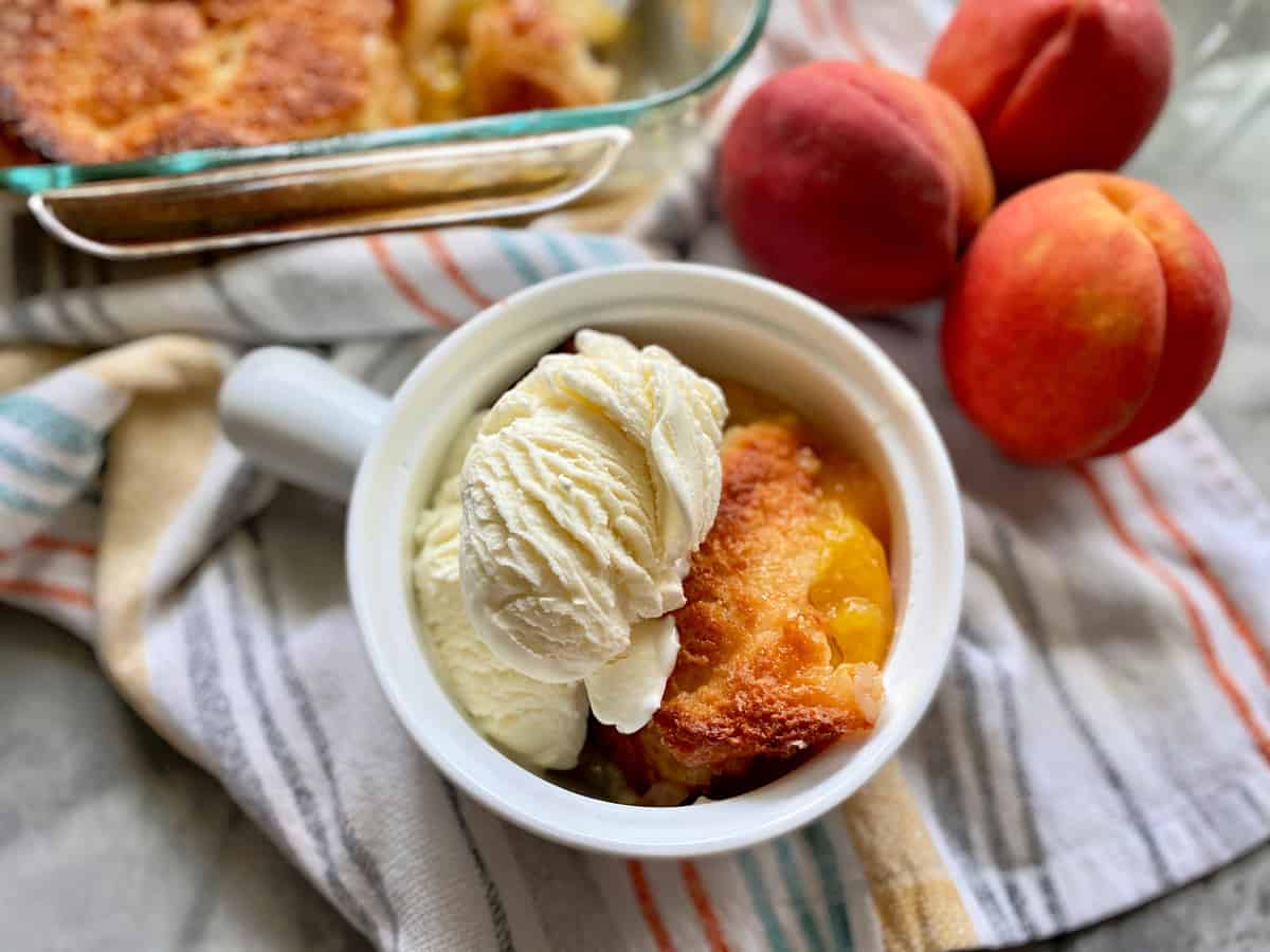 Top view of a white crock with handle filled with peach cobbler and vanilla ice cream sitting on a towel with fresh peaches on the side.