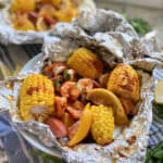 Close up of one foil packet opened with corn, lemon, sausage, and shrimp in a white bowl.