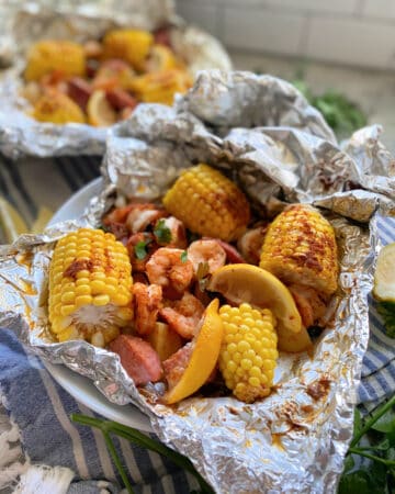 Close up of one foil packet opened with corn, lemon, sausage, and shrimp in a white bowl.