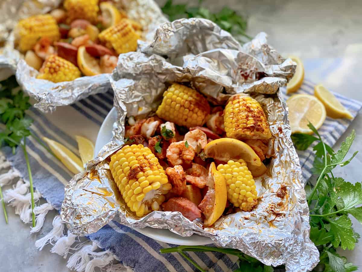 Two foil pouches in a white bowl filled with corn, sausage, shrimp, lemon wedges, and potatoes.