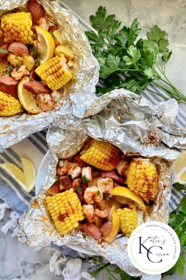 Top view of two foil packets filled with corn, susage, potatoes, shrimp, and lemon wedges.