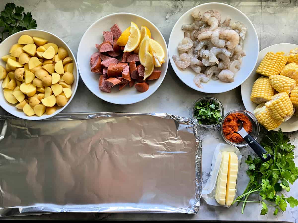 Top view of a bowl of quartered potatoes, diced kielbasa, lemon wedges, shrimp, corn on the cob, parsley, butter, old bay seasoning, and foil on counter.