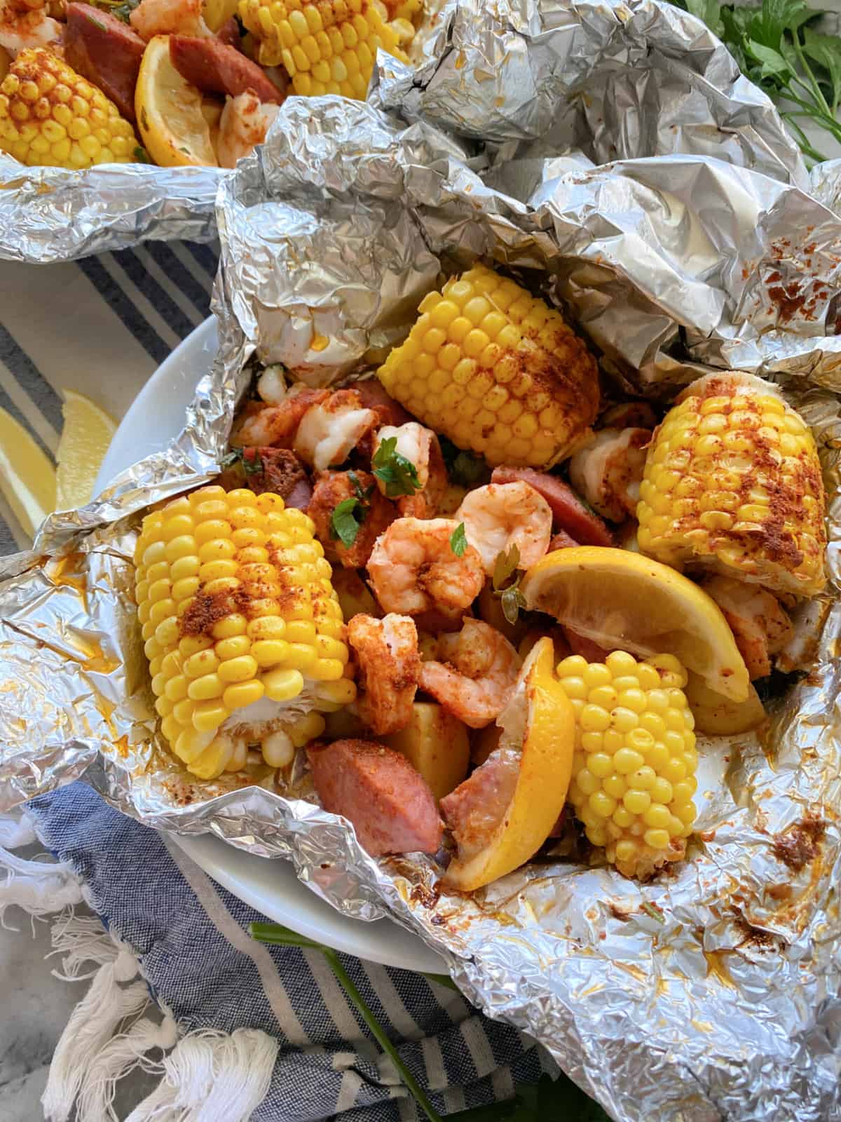 Close up of a white bowl lined with foil with corn on the cob, shrimp, sausage, and lemon wedges.