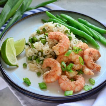 Round white plate with shrimp, rice, green onions and green beans on a plate laying on a cloth.