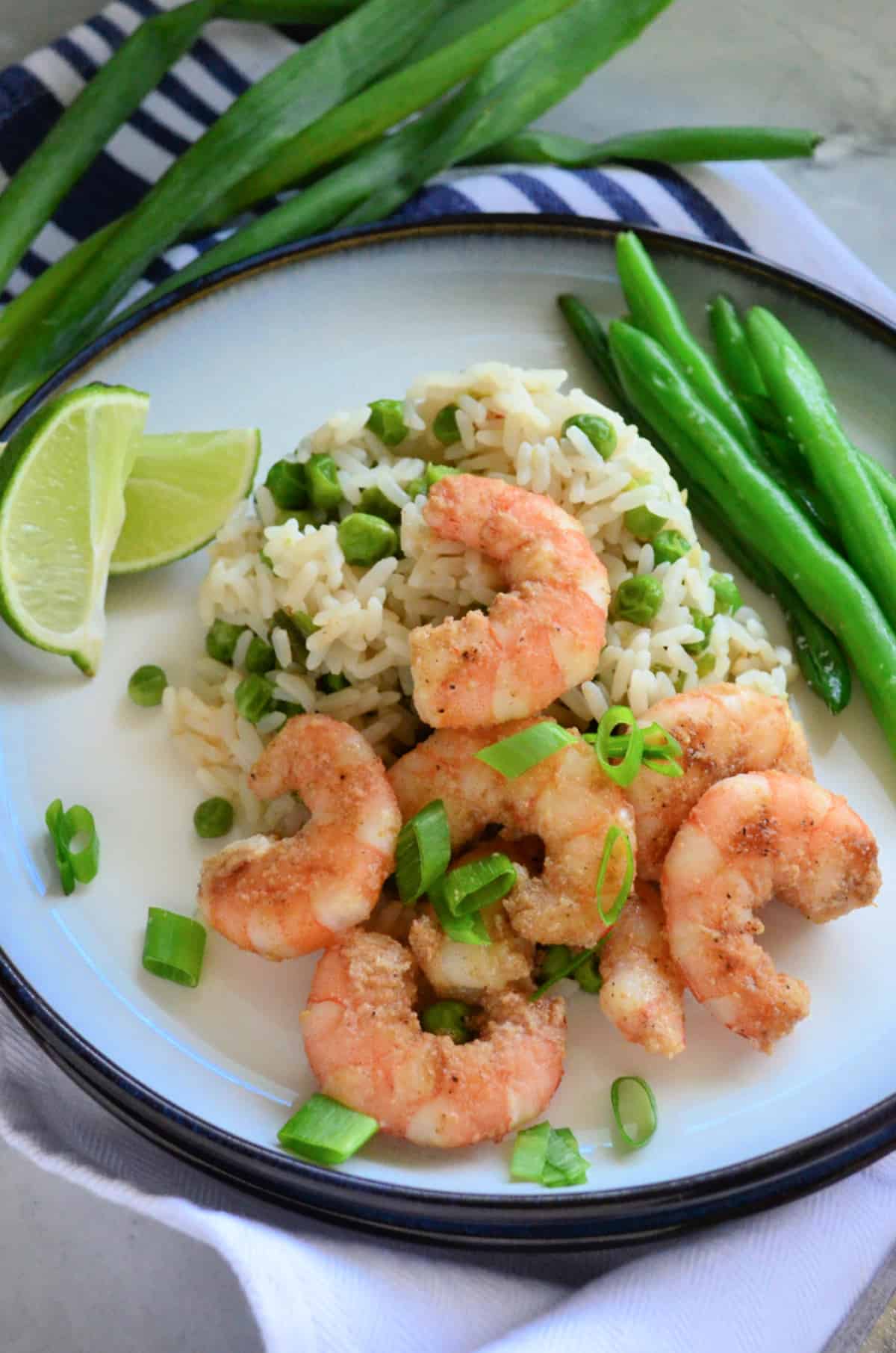 White plate with a blue rim sitting on a counter filled with shrimp, rice, green peas, green onions, and green beans.