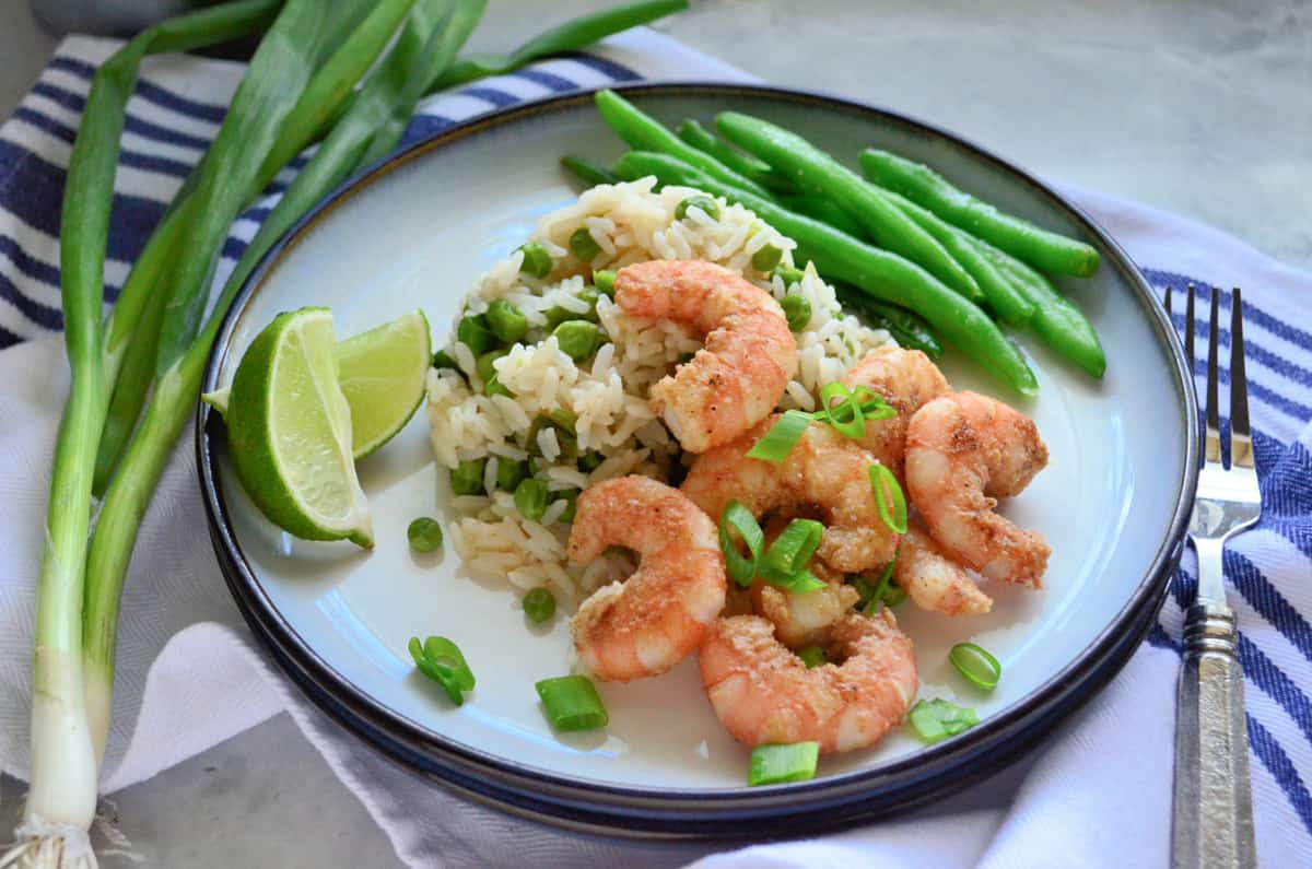 Round white plate with shrimp, rice, green onions and green beans on a plate laying on a cloth.