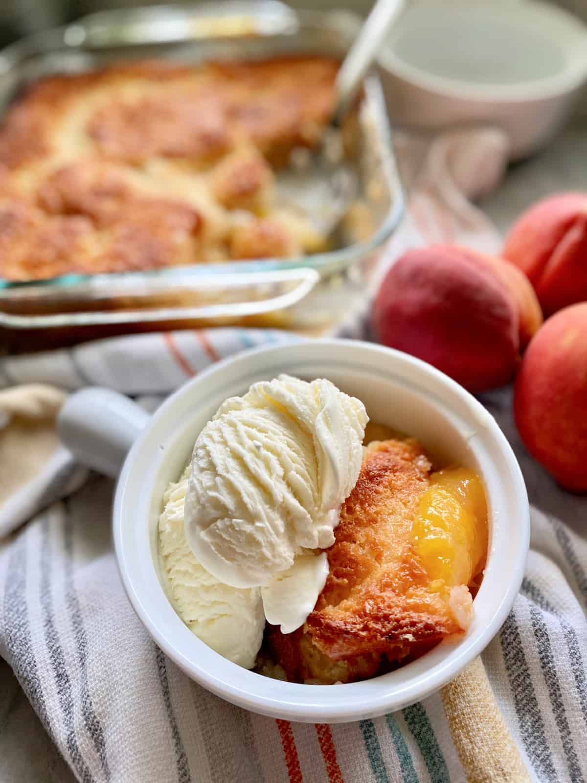 White crock filled with vanile ice cream and peach cobbler on a kitchen towel with peaches and a peach cobbler in the background.