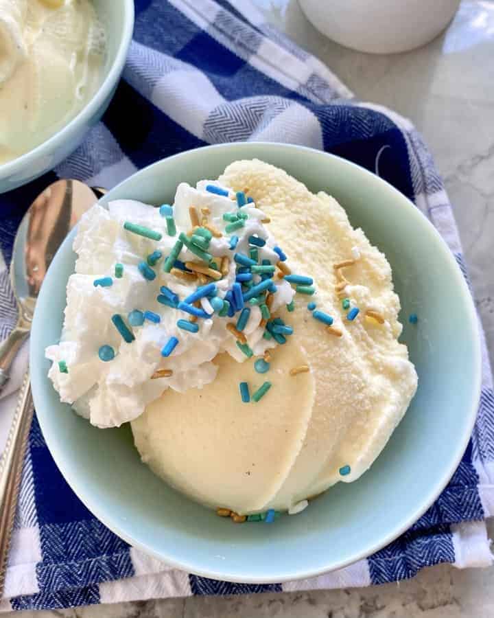 Top view of a light blue bowl filled with vanilla ice cream, whipped cream, and sprinkles with spoons on the side.