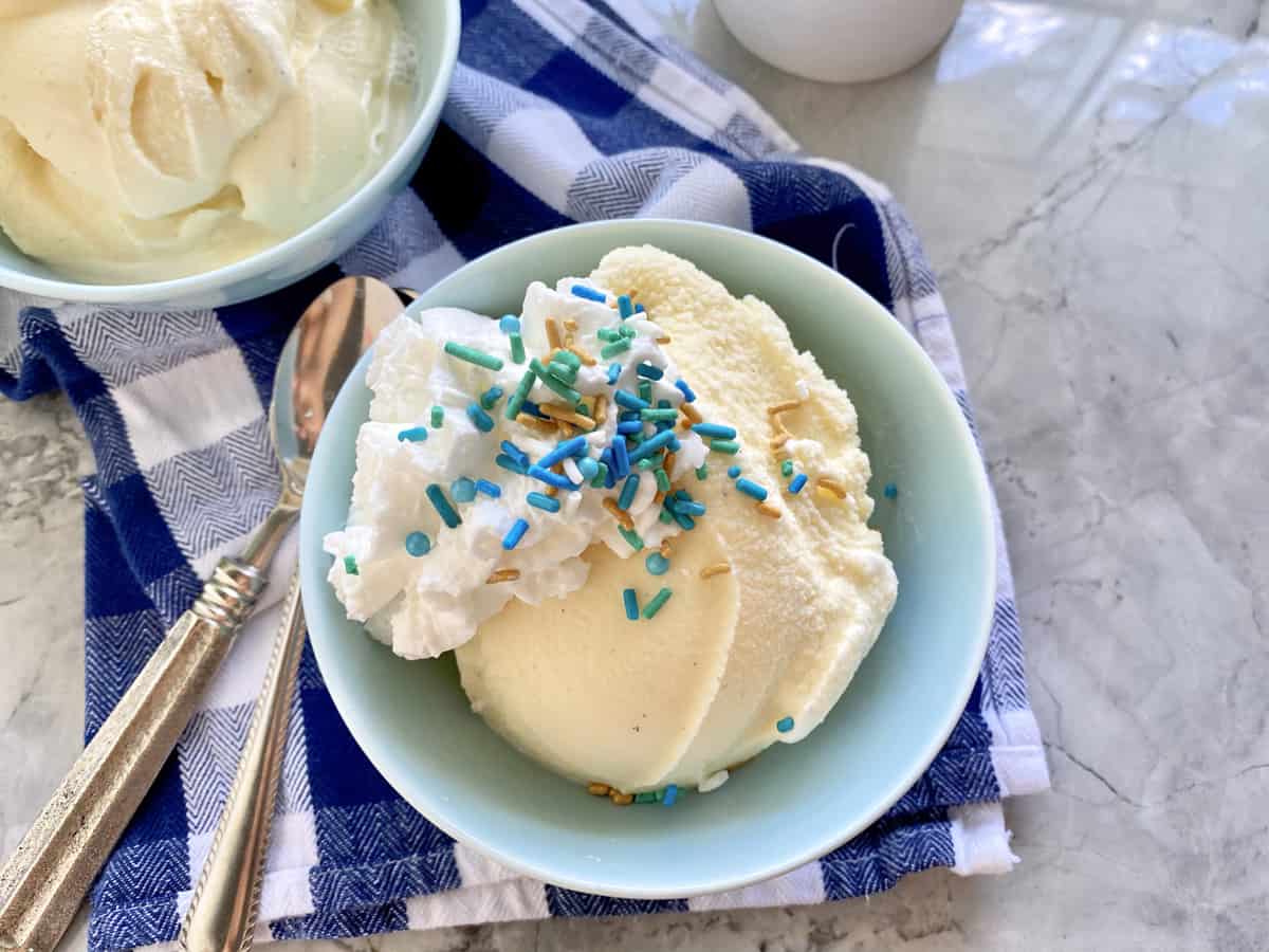 Top view of a light blue bowl filled with vanilla ice cream, whipped cream, and sprinkles with spoons on the side.