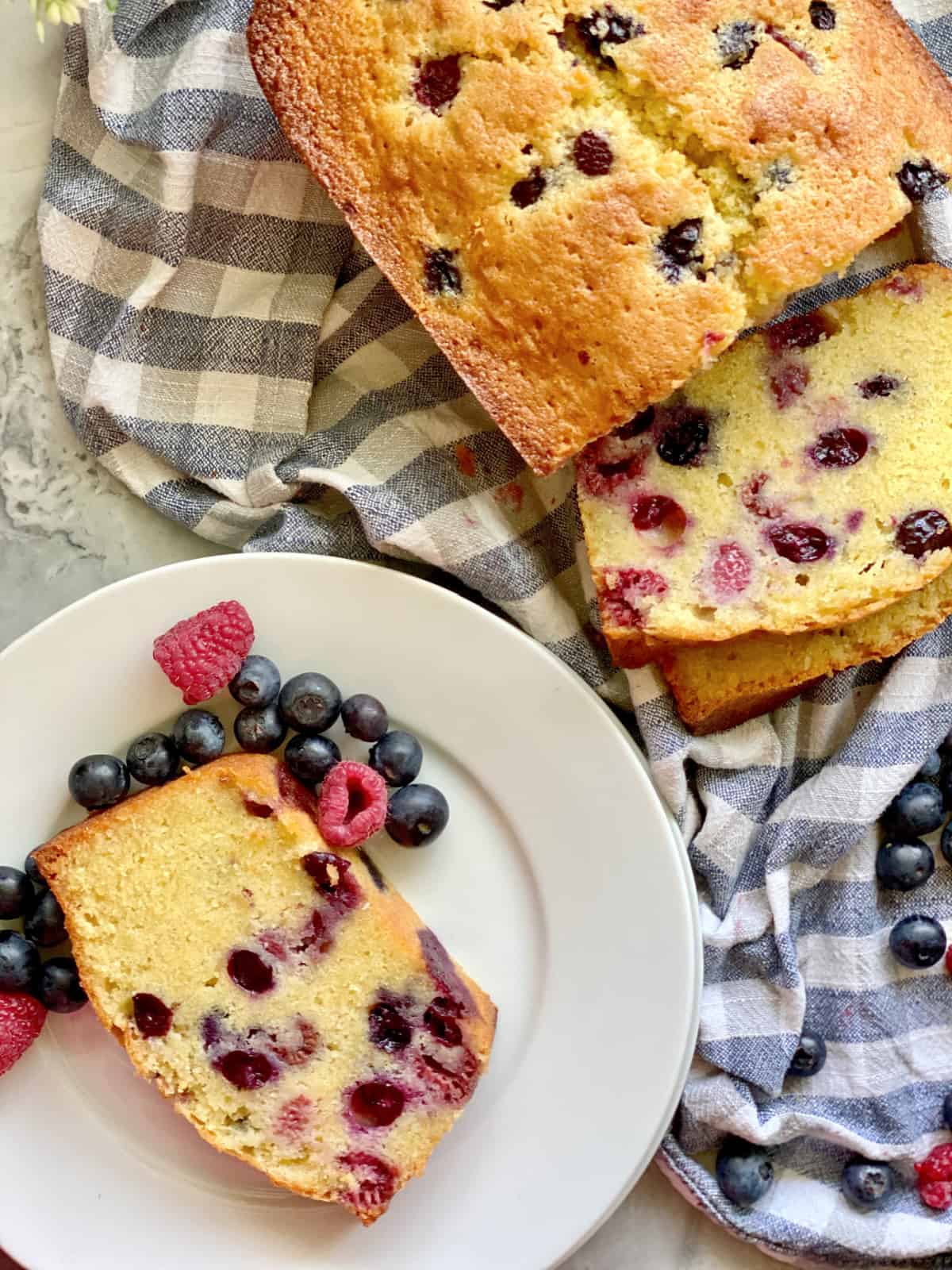 Top view of a sliced loaf cake with a white plate with a sliced berry cake with fresh berries.