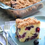 Blue square plate with a slice of Blueberry Coffee Cake with fresh blueberries and a glass baking dish filled with cake in the background.