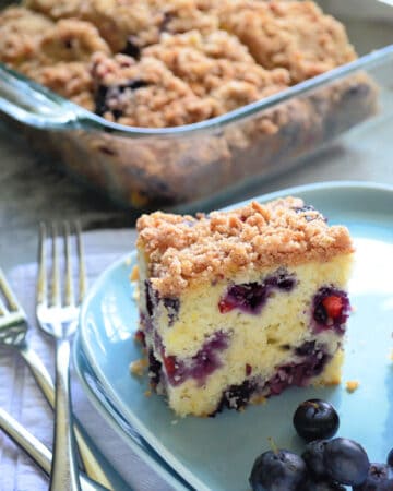 Blue square plate with a slice of Blueberry Coffee Cake with fresh blueberries and a glass baking dish filled with cake in the background.