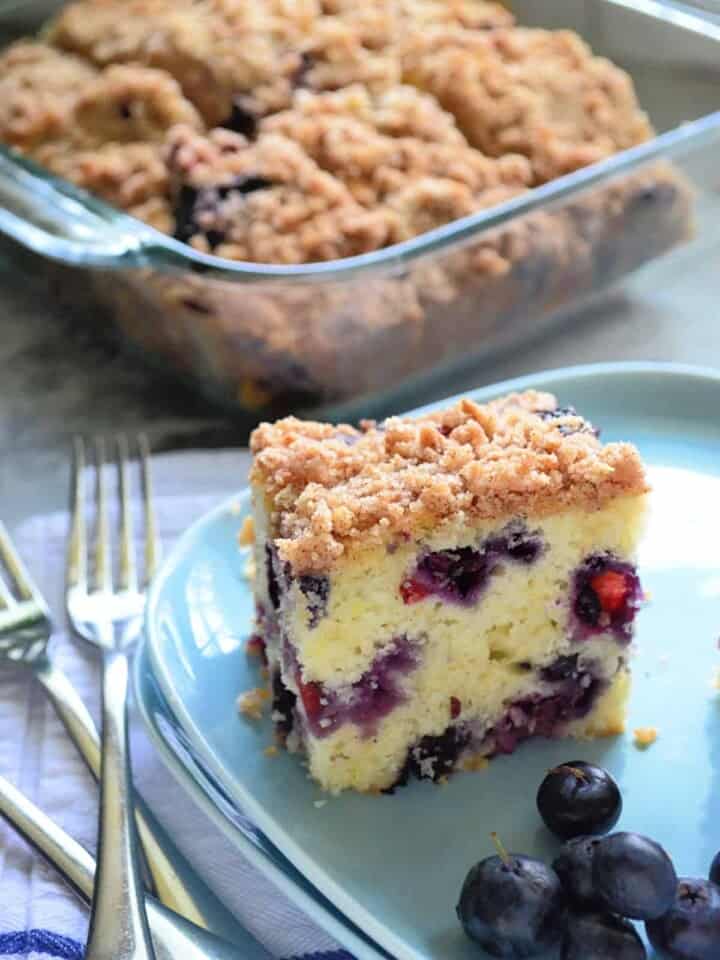 Blue square plate with a slice of Blueberry Coffee Cake with fresh blueberries and a glass baking dish filled with cake in the background.