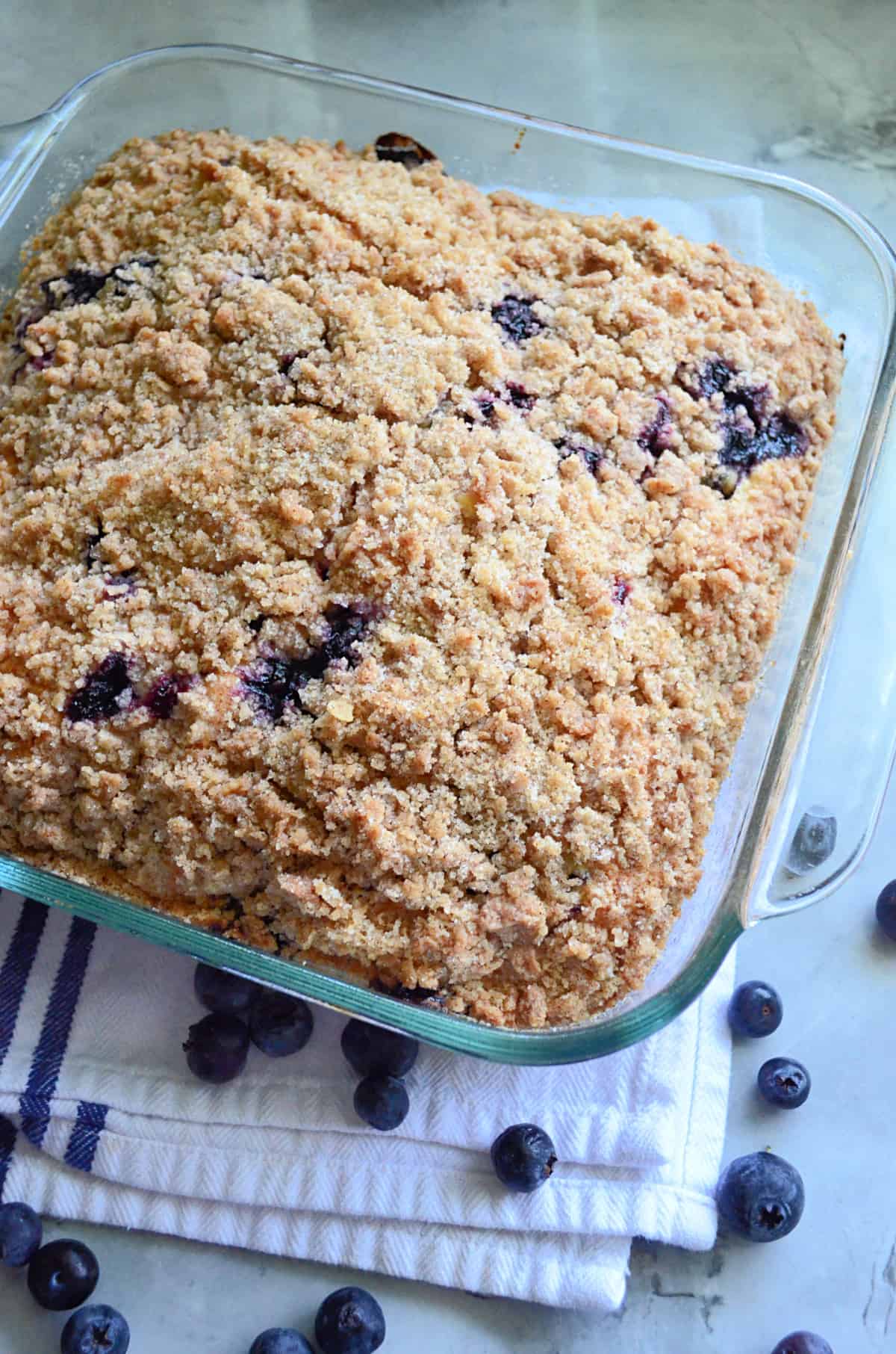 Square glass baking dish filled with a blueberry cake with blueberries on the counter around it.