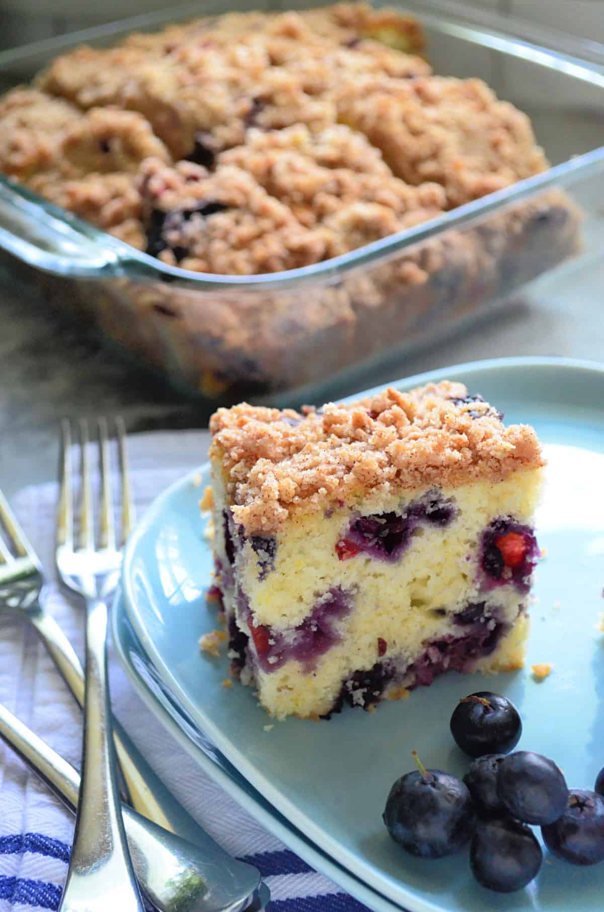 Blue square plate with a slice of Blueberry Coffee Cake with fresh blueberries and a glass baking dish filled with cake in the background.