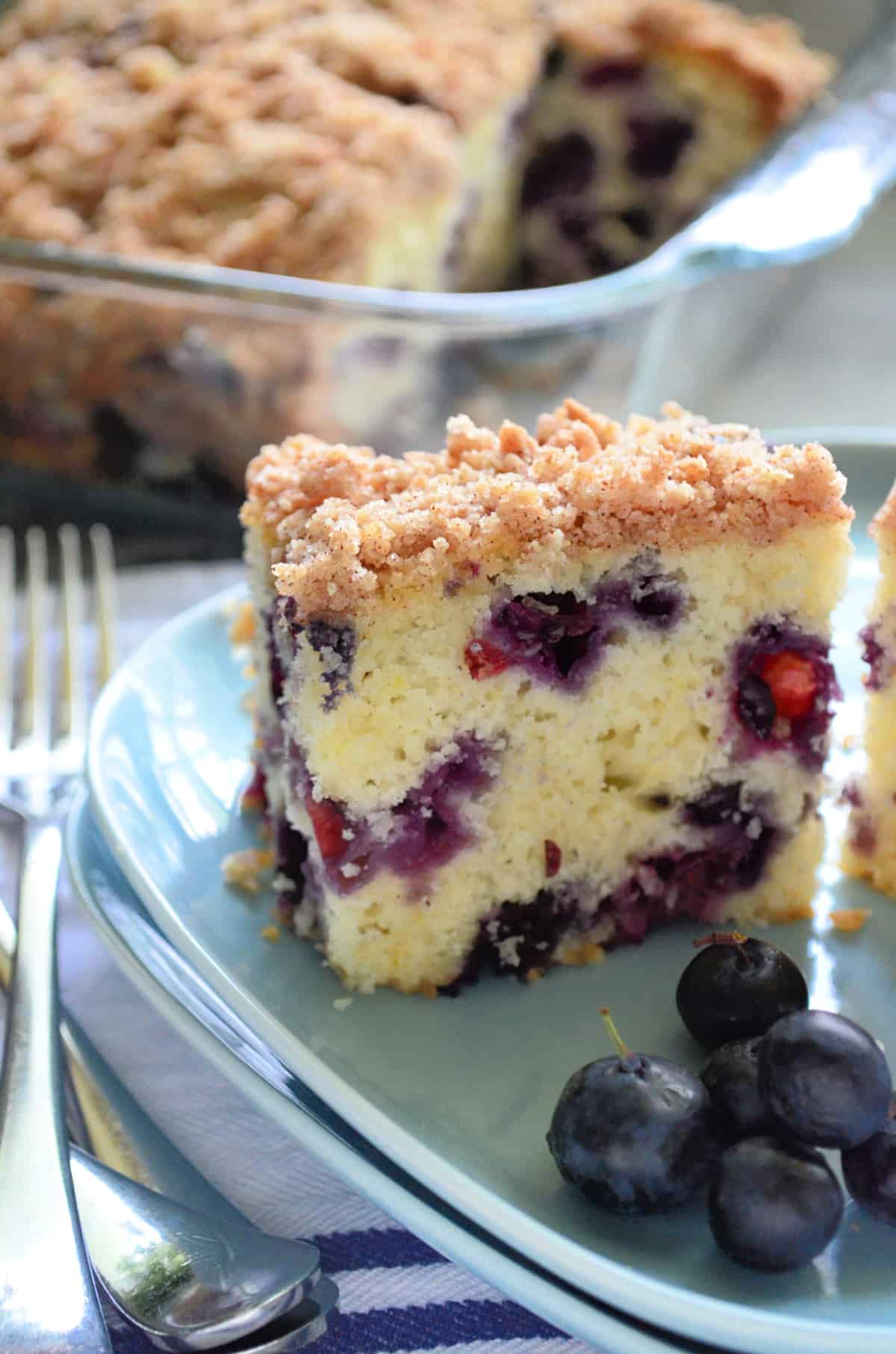 Close up of a slice of Blueberry Coffee Cake on a blue square plate with forks and blueberries on the side.