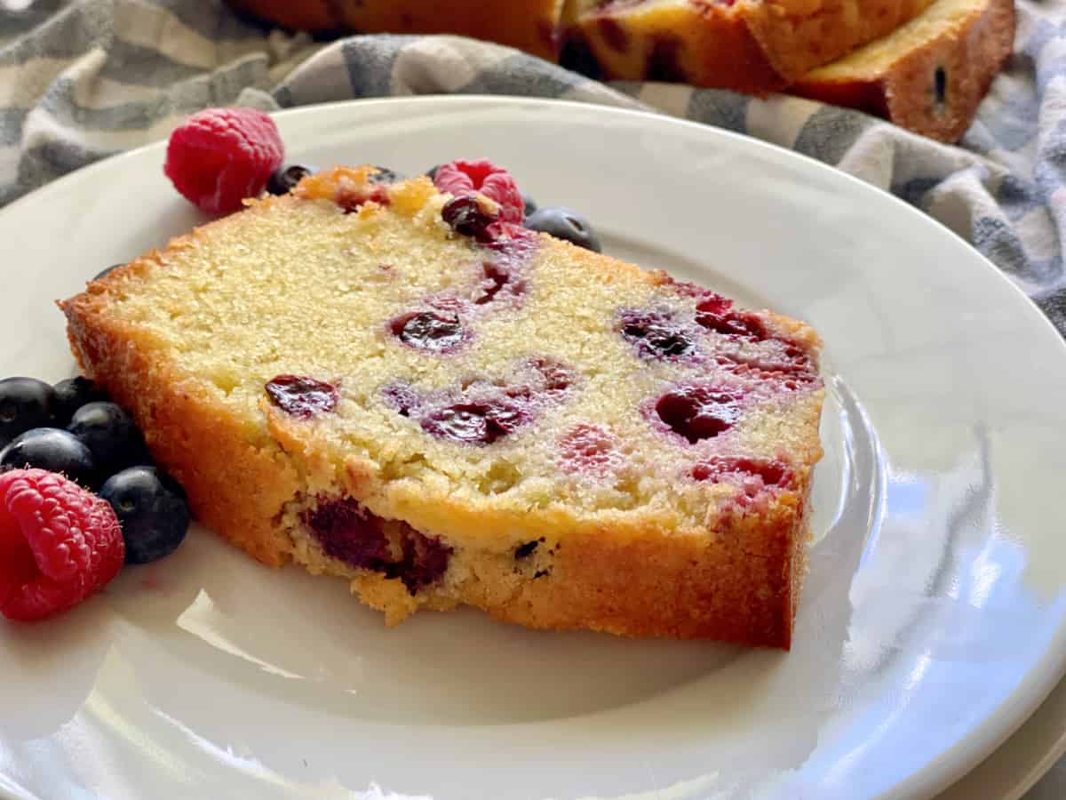 White plate with a slice of Berry Cornmeal Pound Cake with fresh berries.