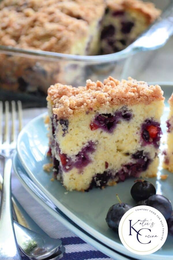 Close up of a slice of Blueberry Buckle with fresh blueberries with a logo on the right corner.