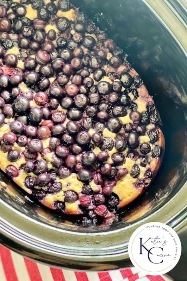 Top view of a slow cooker filled with cooked bluberries and cake with a log on the right corner.