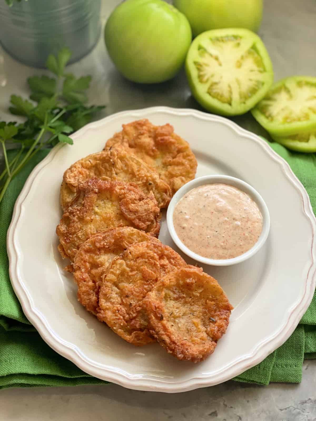 Six golden Fried Green Tomatoes on a white plate with siced green tomatoes on the counter.