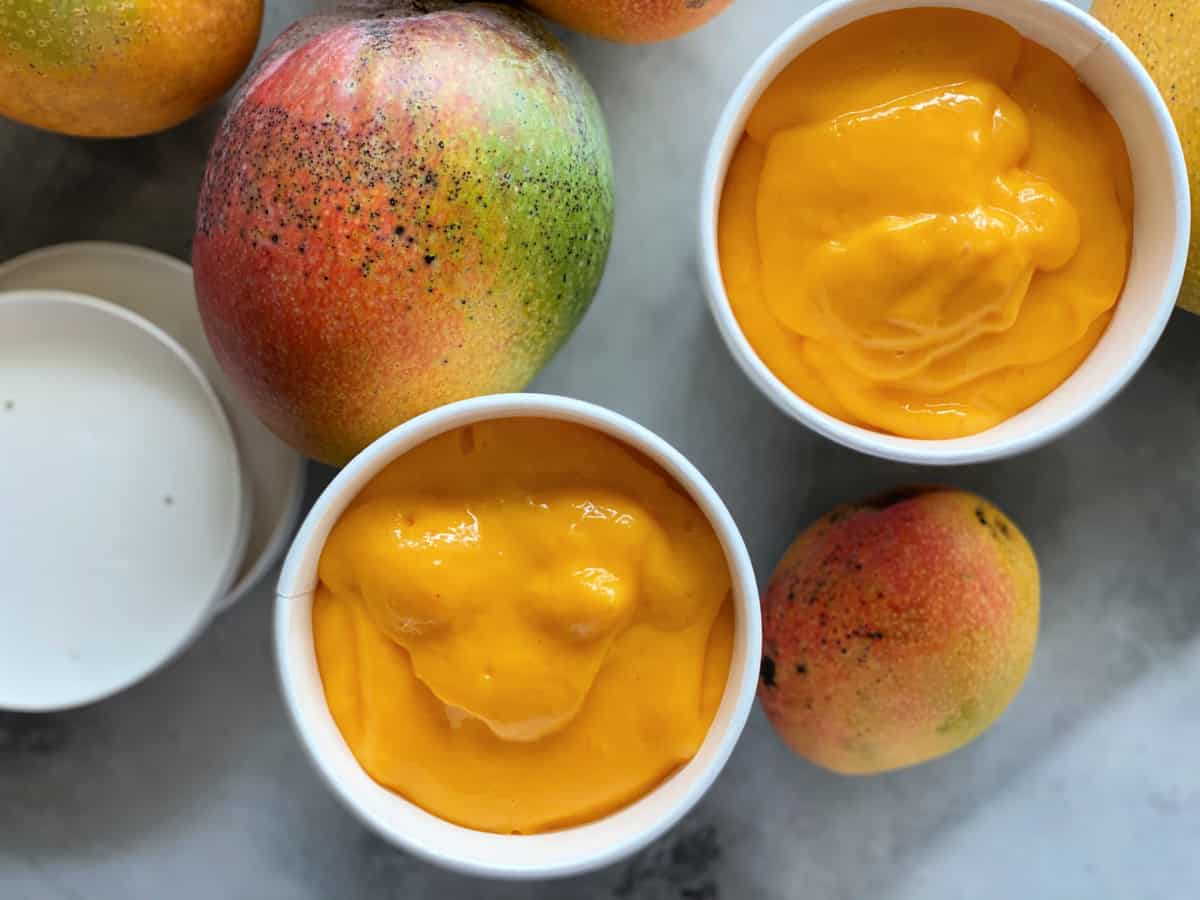 Top view of two white containers filled with Mango Frozen Yogurt and fresh mangoes on the counter.