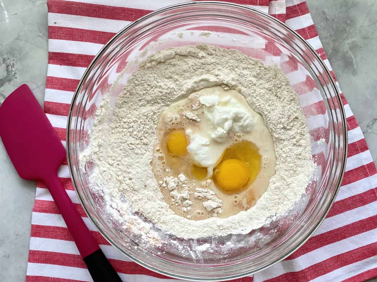 Top view of a glass bowl filled with flour, milk, sour cream, and eggs on a red and white striped cloth.