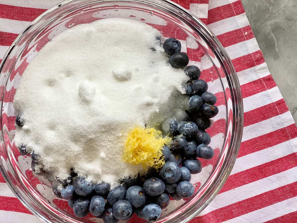 Top view of a glass bowl filled with blueberries, lemon zest, lemon juice, and sugar on a red and white striped cloth.