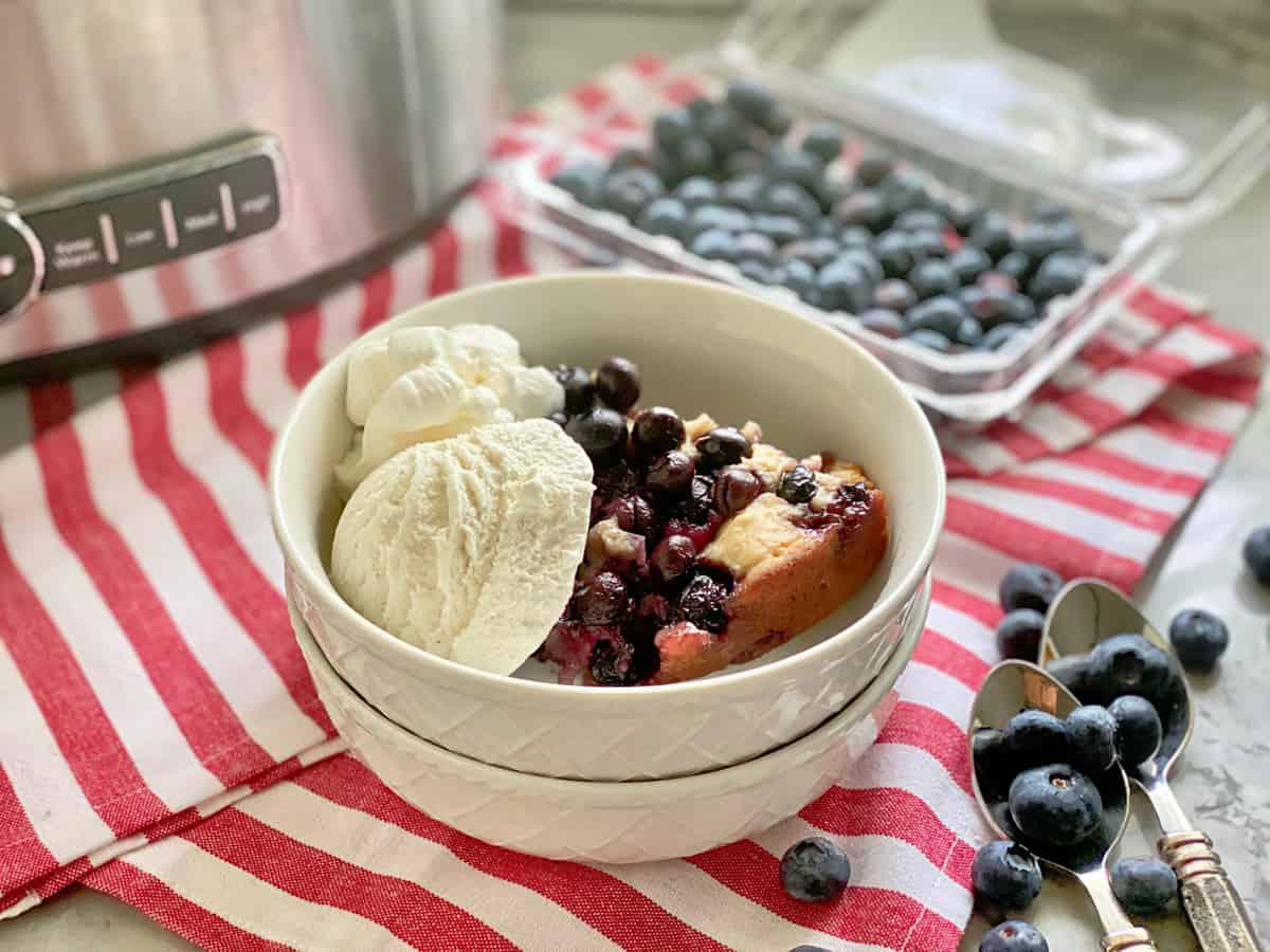 Two stacked white bowls filled with blueberry cobbler with vanilla ice cream with slow cooker in background.