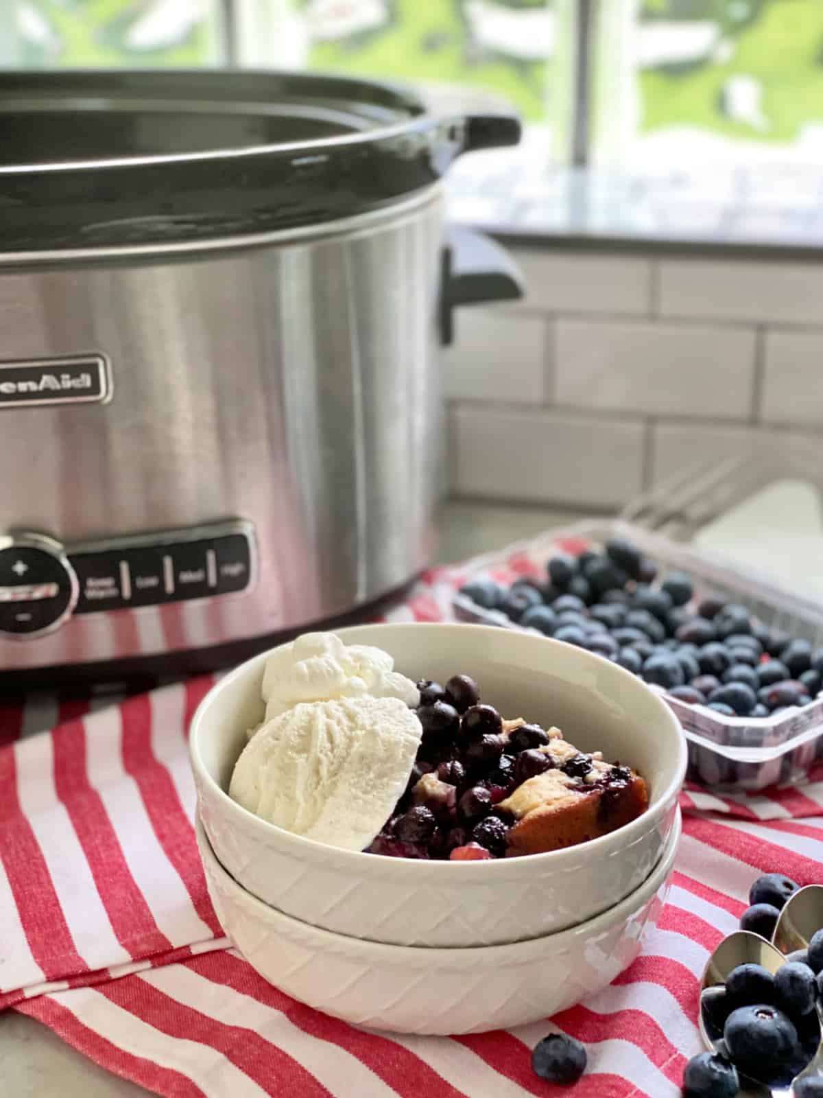 Two white bowls stacked with blueberry cobbler and vanilla ice cream with a stainless steel slow cooker in background.