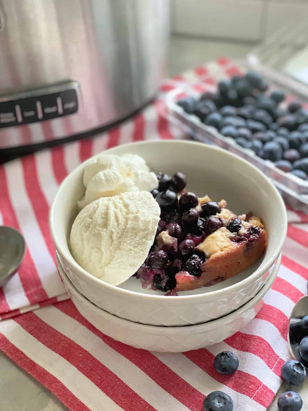 Close up of two stacked white bowls with blueberry cobbler and vanilla ice on a red and white striped cloth.