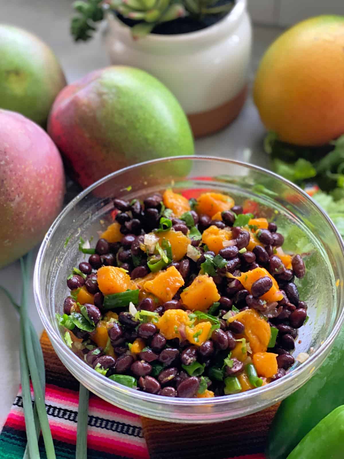 Glass bowl filled with mangos, black beans, and green onion on a multi colored cloth with mangoes in the background.