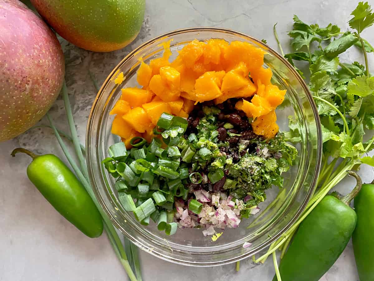 Top view of a glass bowl filled with diced mango, green onions, red onion, cilantro, seasoning, and black beans.